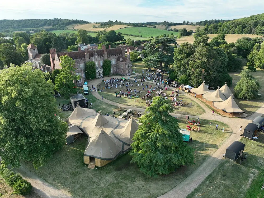 Aerial view of a historic manor complex with numerous beige canvas tents arranged around a central lawn and crowd gathered for an outdoor festival.